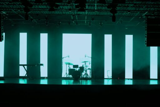 An empty stage with a drum kit and a keyboard in front of tall lit panels.