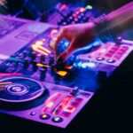 A hand on a lit DJ controller in purple and orange light, with motion blur.