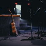 An acoustic guitar and microphone stands on a quiet, dimly lit stage.