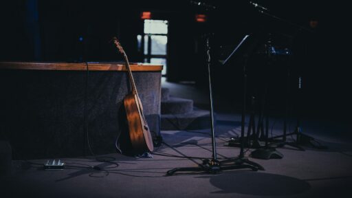An acoustic guitar and microphone stands on a quiet, dimly lit stage.