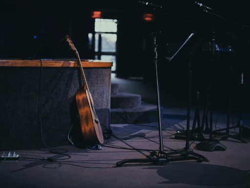 An acoustic guitar and microphone stands on a quiet, dimly lit stage.
