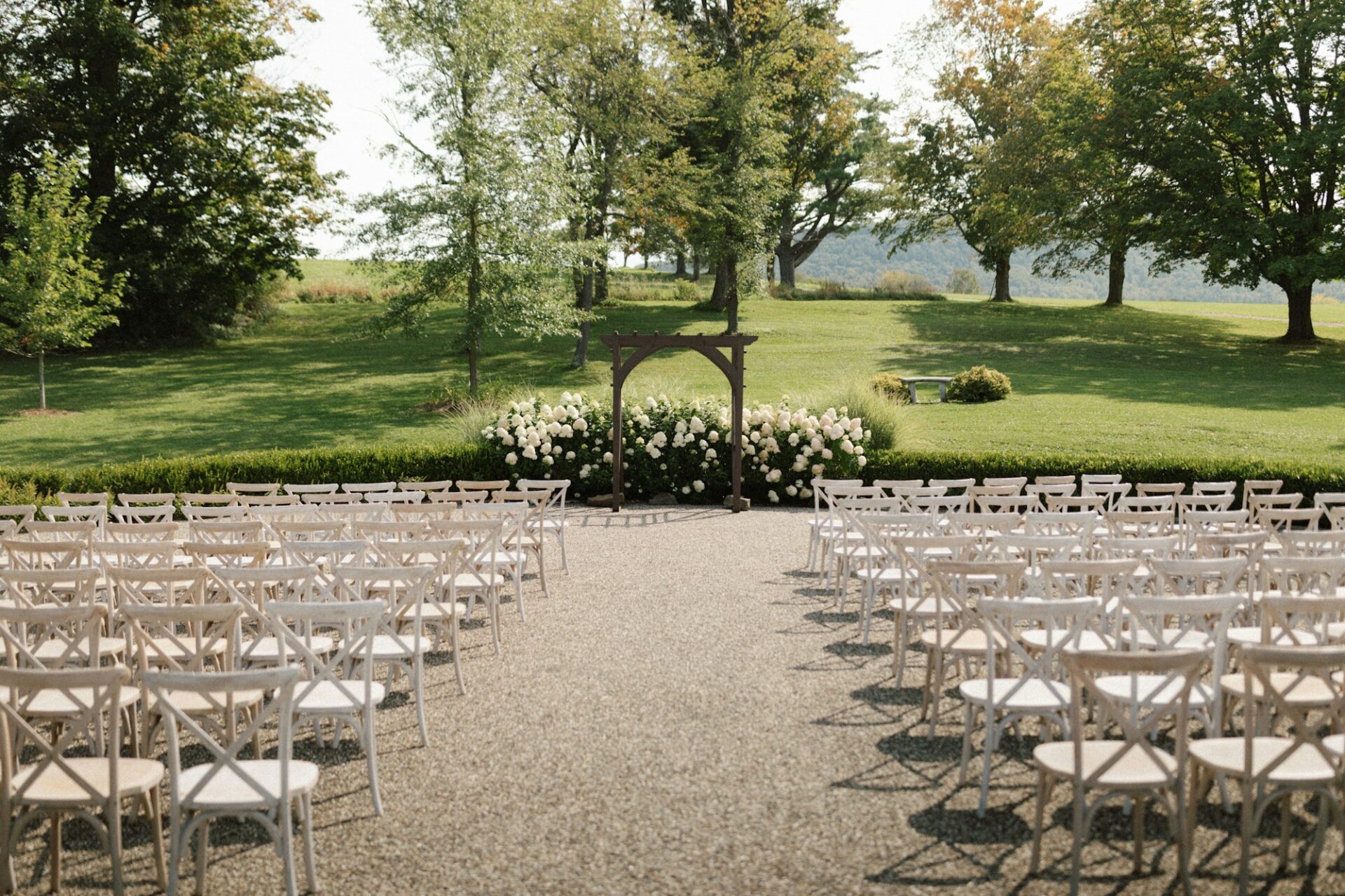 Rows of white wooden chairs either side of an aisle leading to a wooden arch with white flowers, on a lawn.