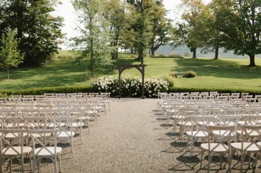 Rows of white wooden chairs either side of an aisle leading to a wooden arch with white flowers, on a lawn.