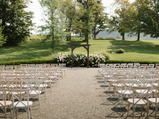 Rows of white wooden chairs either side of an aisle leading to a wooden arch with white flowers, on a lawn.