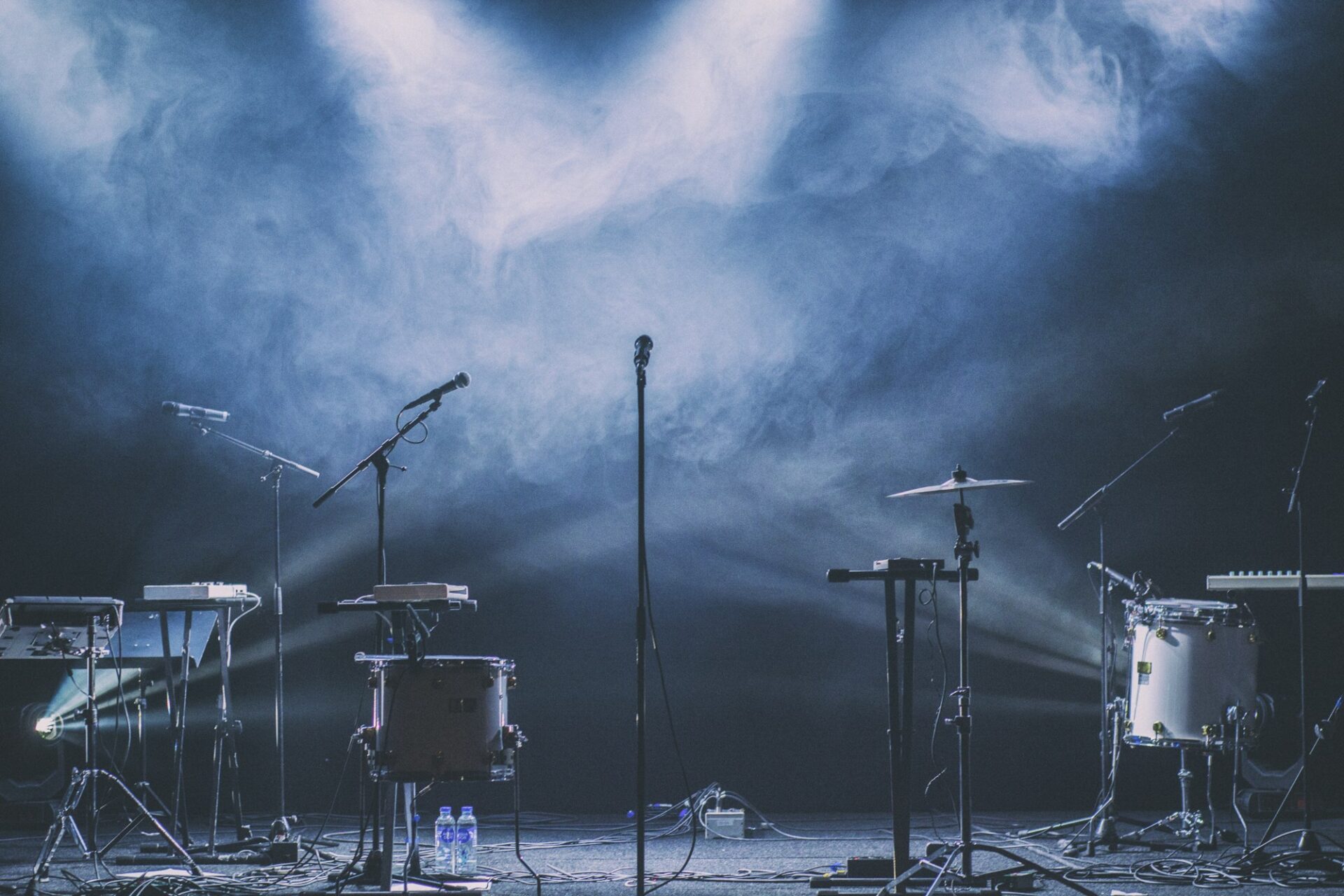 Drums, keyboards and microphone stands on an empty stage under blue spotlights and haze.