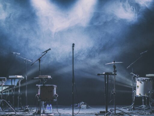 Drums, keyboards and microphone stands on an empty stage under blue spotlights and haze.