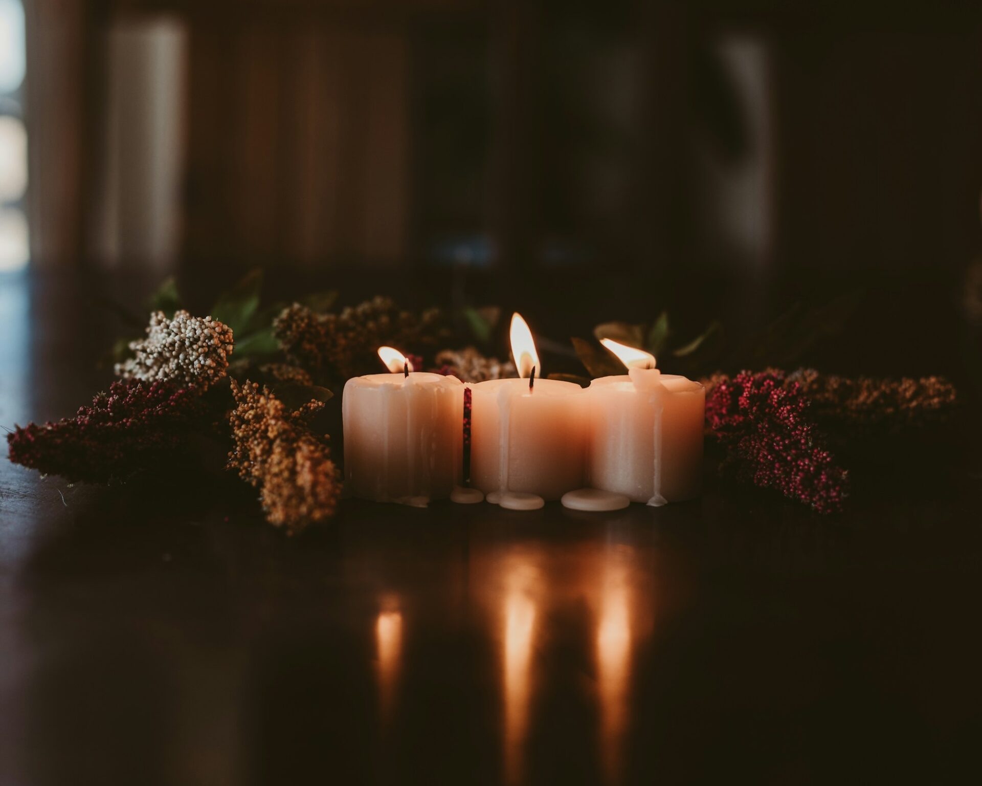 Three lit white candles among dried flowers on a dark wooden table.