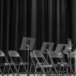 Empty folding chairs and music stands on a stage in front of a dark curtain.