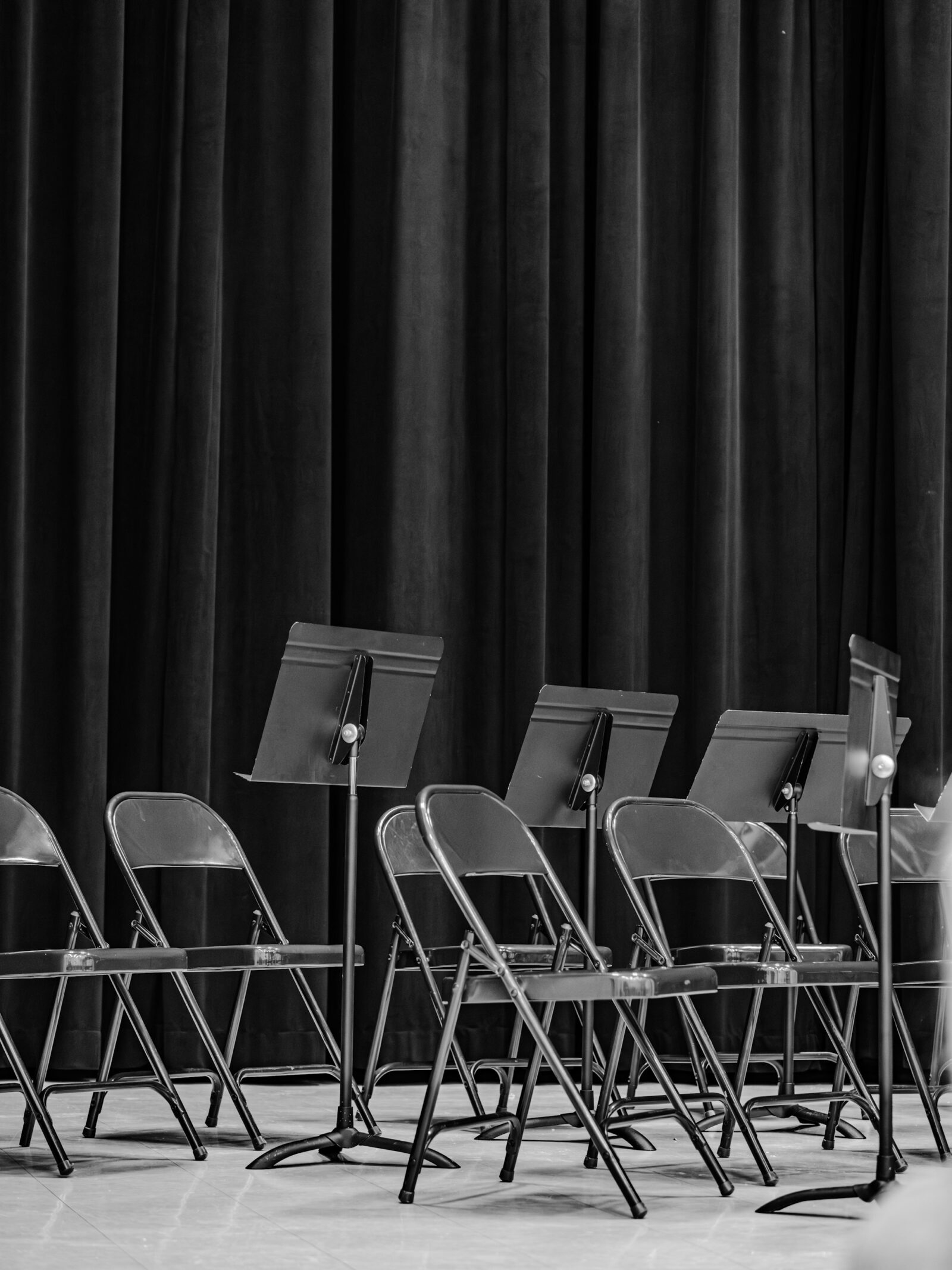 Empty folding chairs and music stands on a stage in front of a dark curtain.