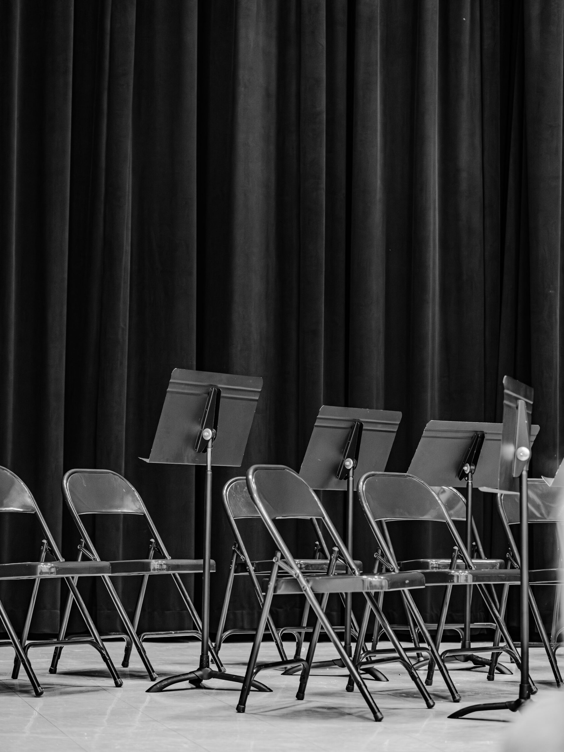 Empty folding chairs and music stands on a stage in front of a dark curtain.