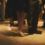 The legs and shoes of dressed-up guests on a lit dance floor, in warm low light.