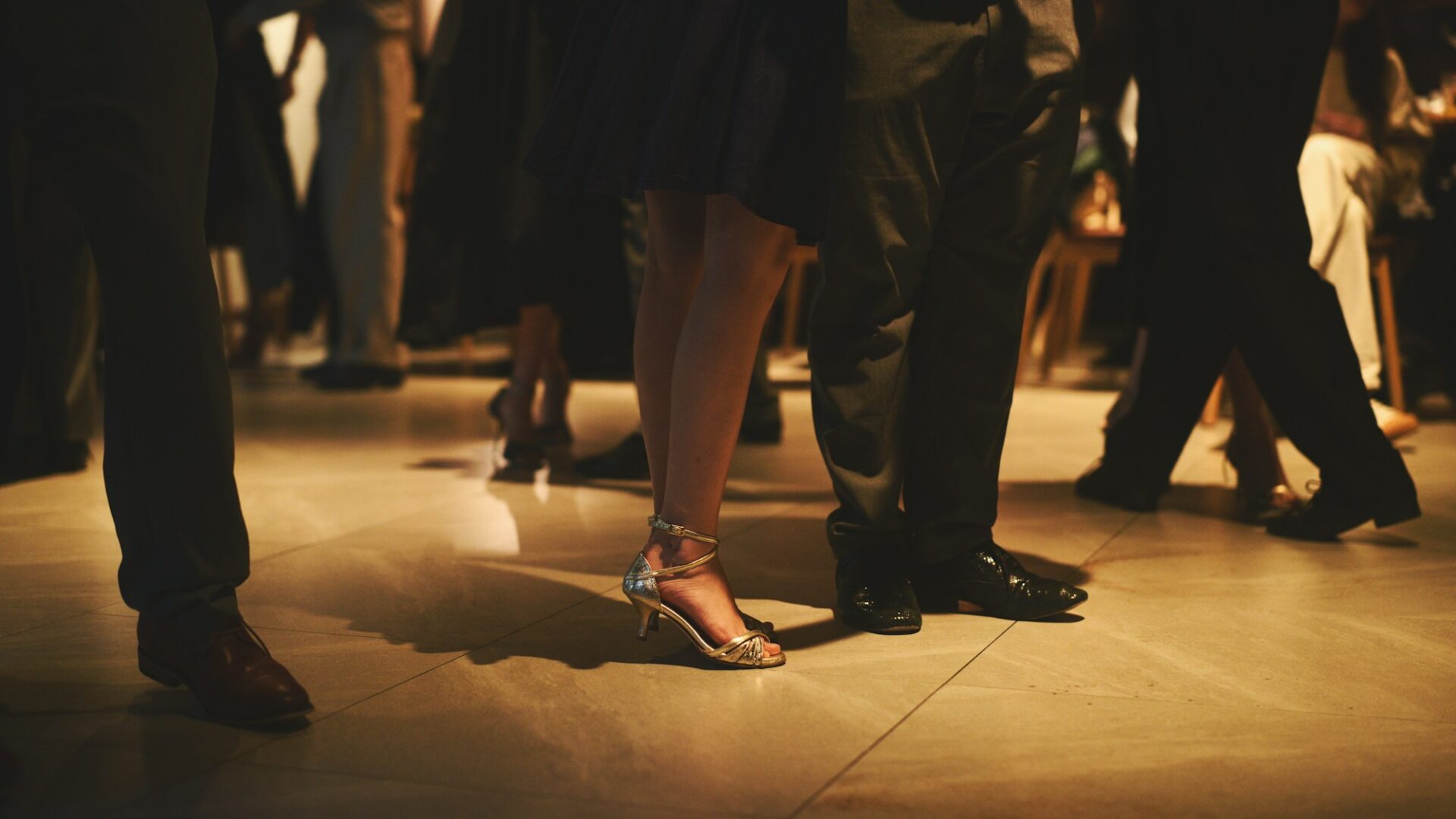 The legs and shoes of dressed-up guests on a lit dance floor, in warm low light.