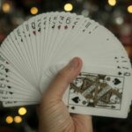 One hand holding a fanned deck of playing cards with the queen of spades showing, warm out-of-focus Christmas tree lights behind.
