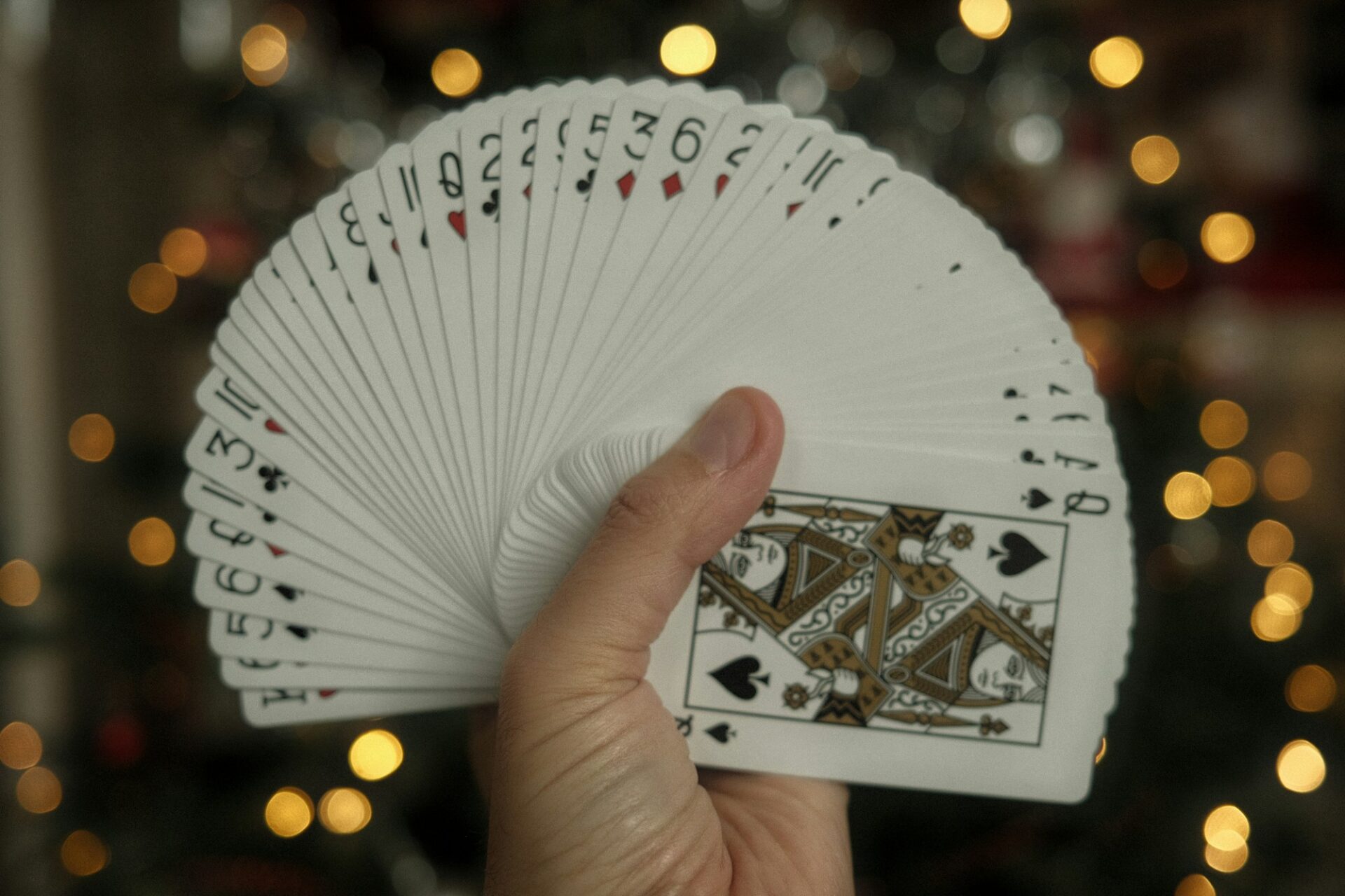 One hand holding a fanned deck of playing cards with the queen of spades showing, warm out-of-focus Christmas tree lights behind.